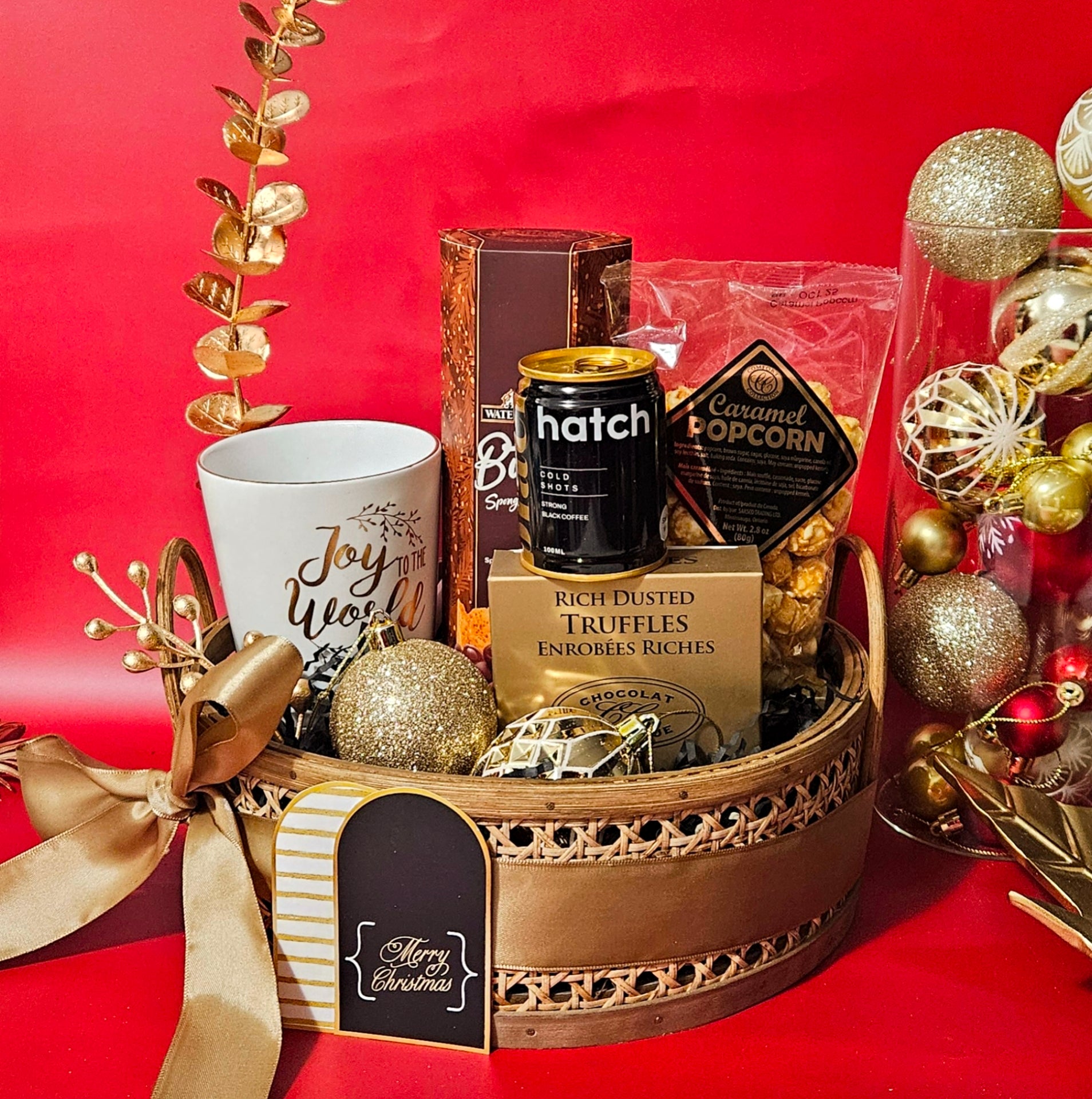 A circular rattan basket containing a gold-engraved coffee mug, a box of buttery toffee, a box of rich truffles, caramel popcorn, a cold coffee shot from Hatch Crafted, and glittering Christmas ornaments, arranged against a red background with golden decorations.