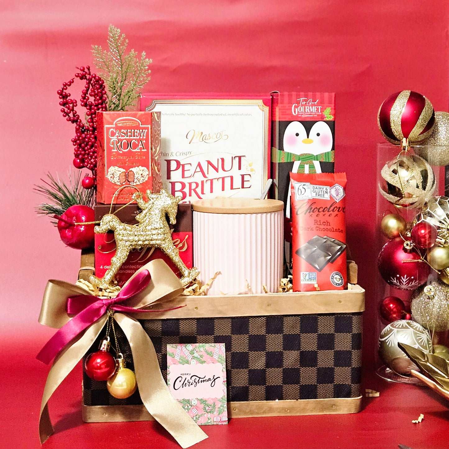 A wicker basket arranged with a red coffee mug, peanut brittle, almond roca, truffles, a sugar cookie, and dark chocolate, adorned with gold and pink decorative ribbons, and a rocking horse tree ornament.