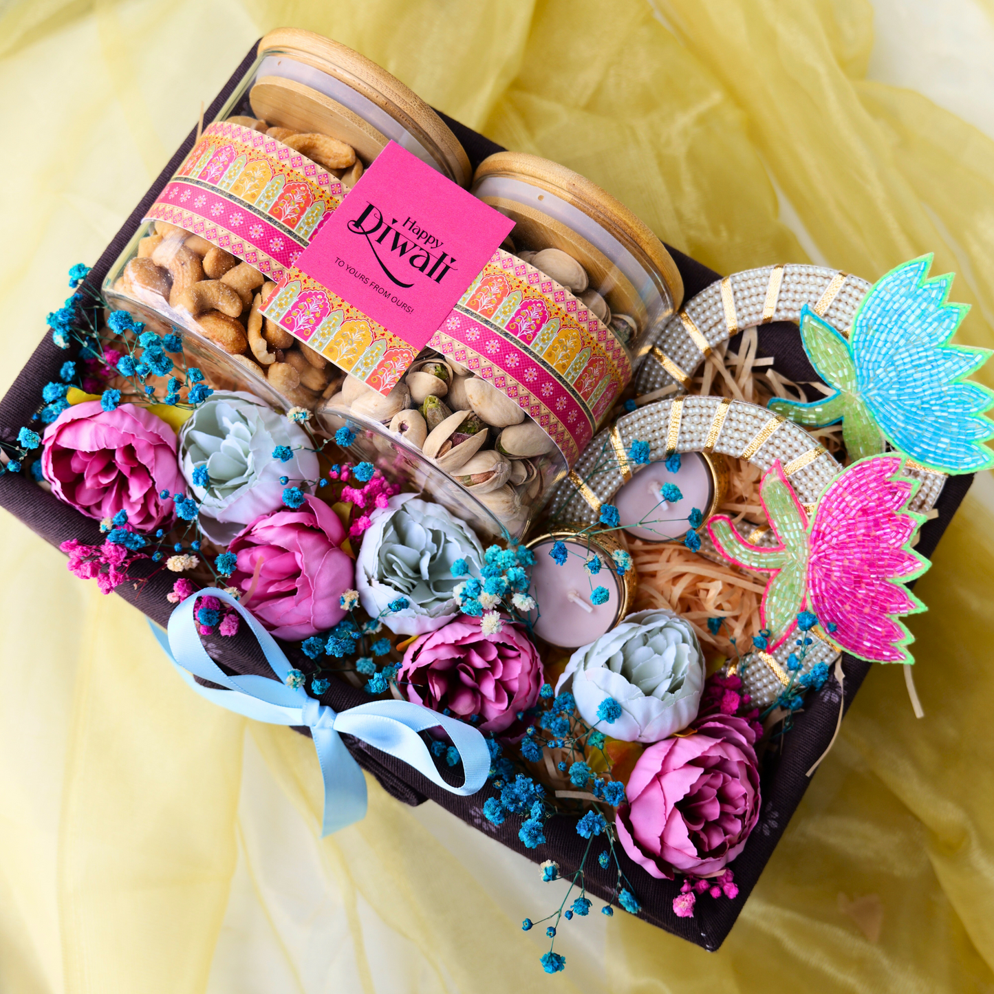 Decorative box with Diwali-themed items including flowers, ribbons, and a 'Happy Diwali' card on a yellow fabric background.