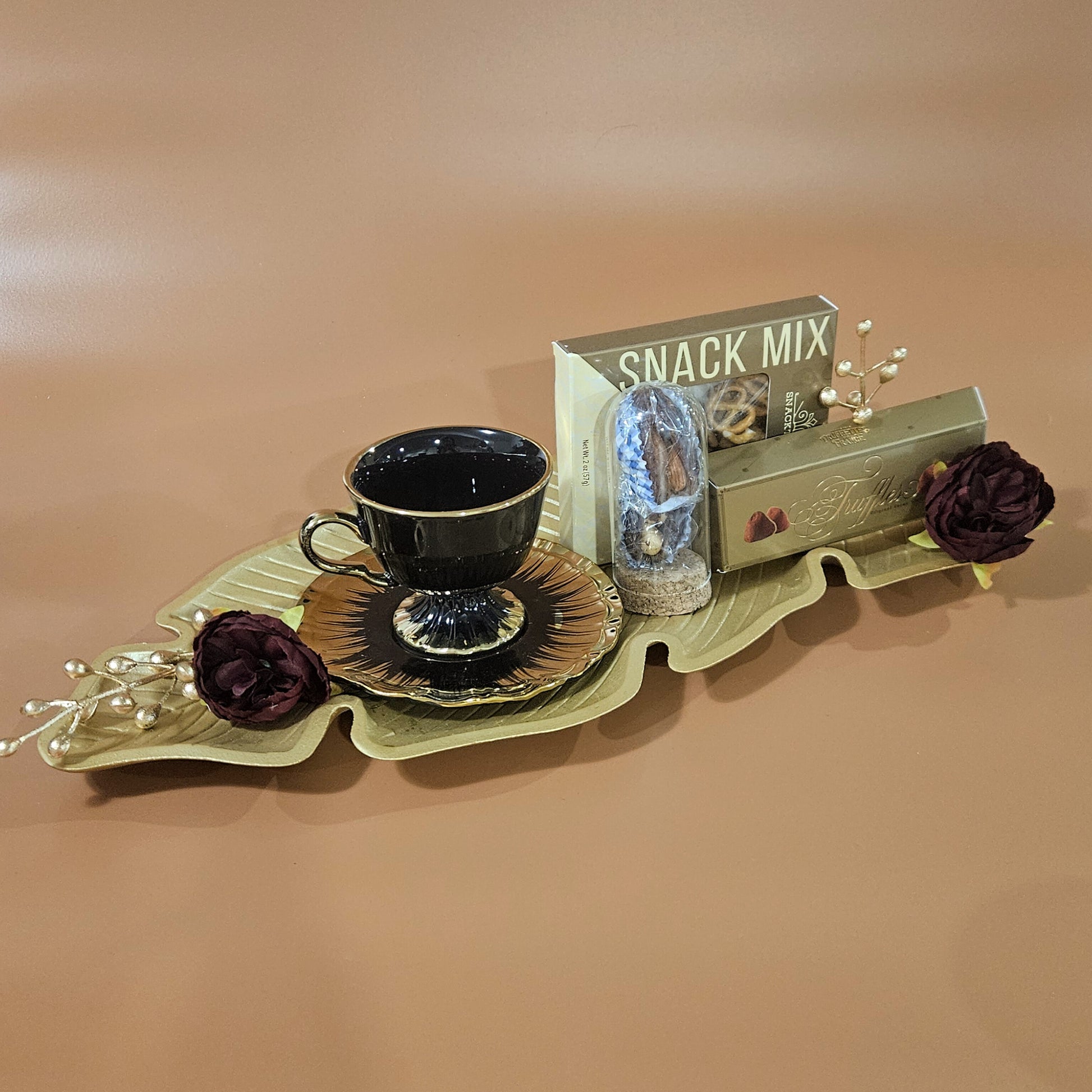 A gold leaf platter with a black cup and saucer, a snack mix box, and some decorative flowers on a brown background.
