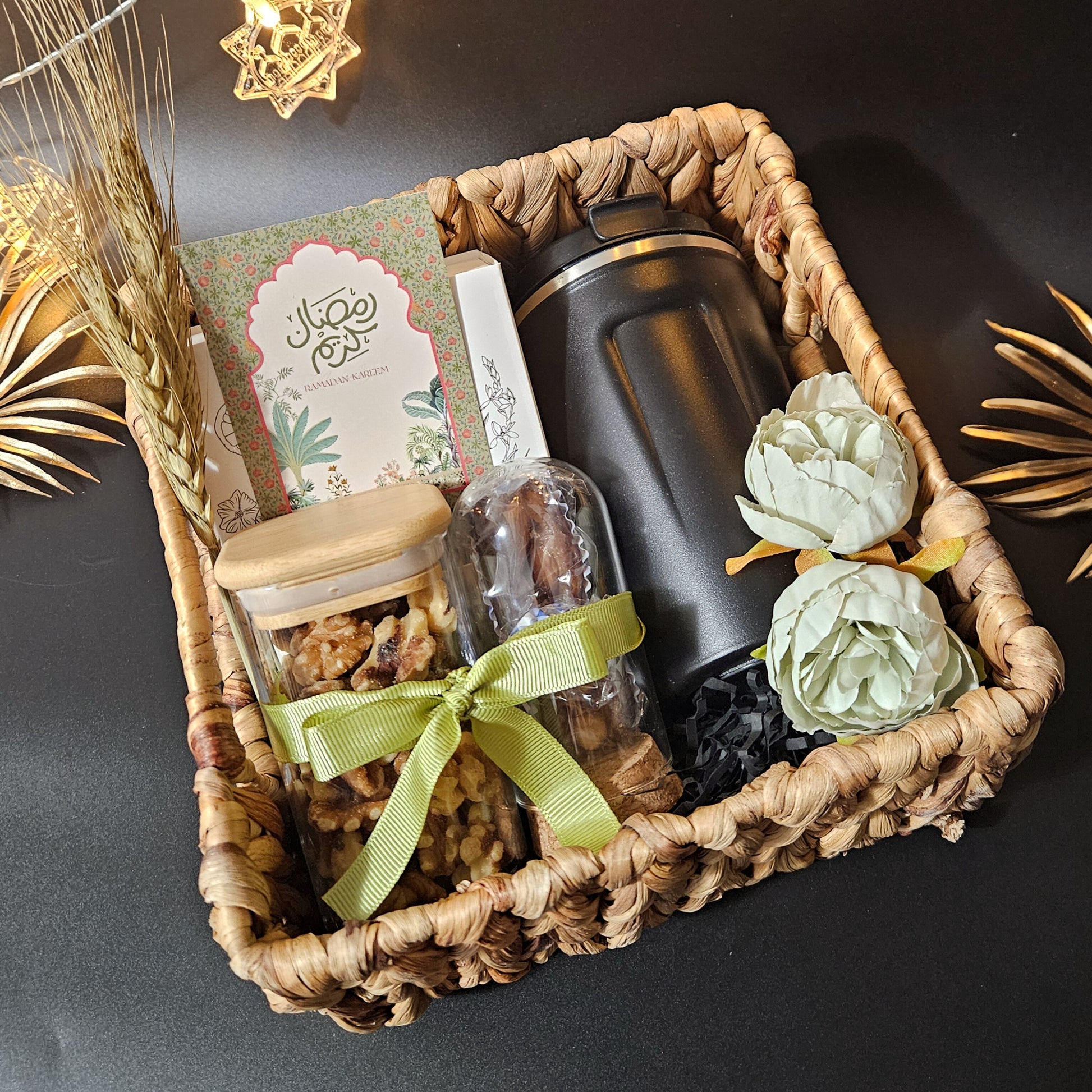 A square cane basket gift hamper arranged with a hot/cold flask, car diffuser, miniature cloche with dates, and dry fruits jar.