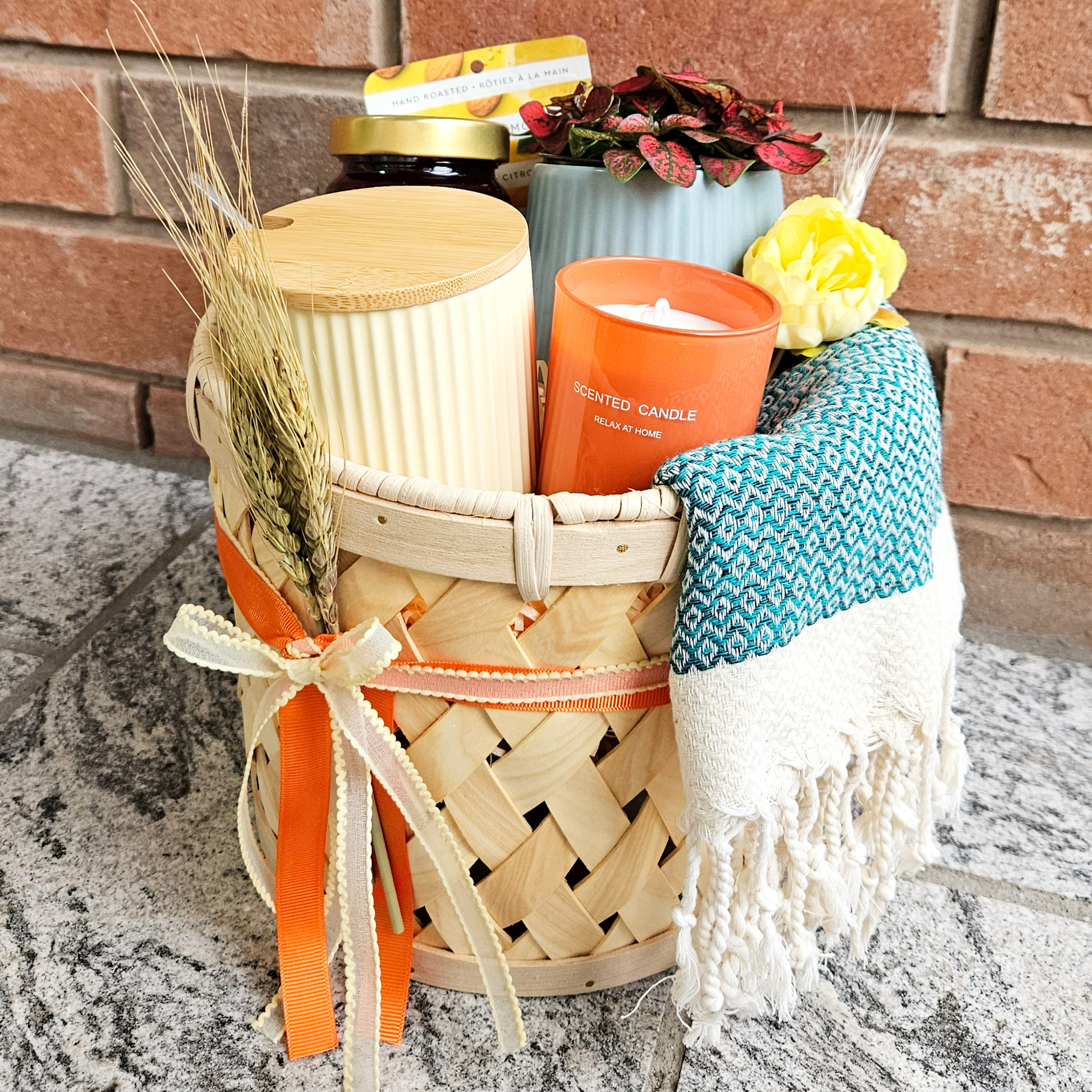 A wicker gift basket containing a soy wax candle, a pair of cotton kitchen cloths, a pastel coffee mug, and a wholegrain mustard plaster, adorned with a decorative scarf and a white throw blanket.