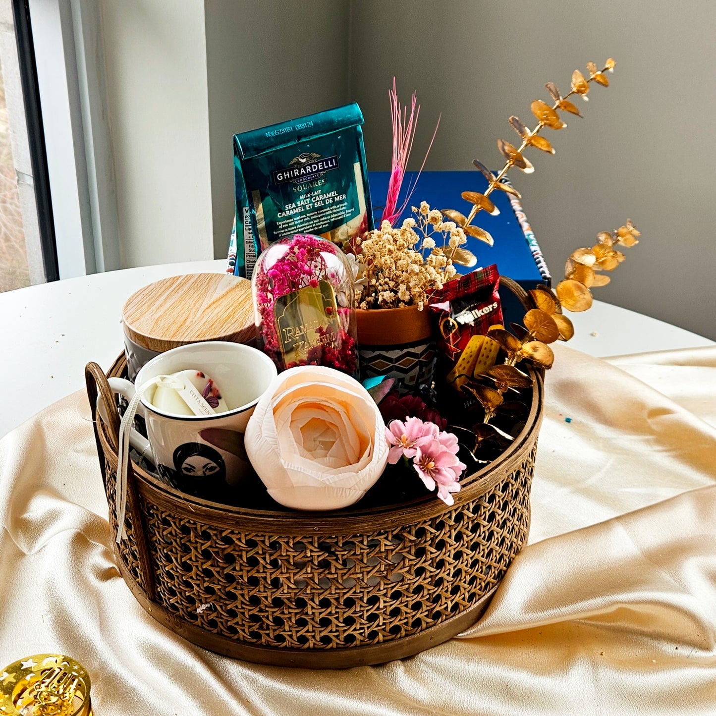 A rattan basket gift hamper containing a variety of items including a printed vase, a black tea cup, a tin with cookies, and a glass cloche with 'Eid Mubarak' plating, all beautifully arranged on a table with a golden yellow tablecloth.