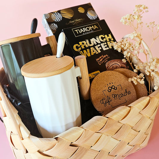 A gift hamper containing two coffee mugs with wooden handles, two cork coasters, and a pack of cookies, all presented in a wicker basket.