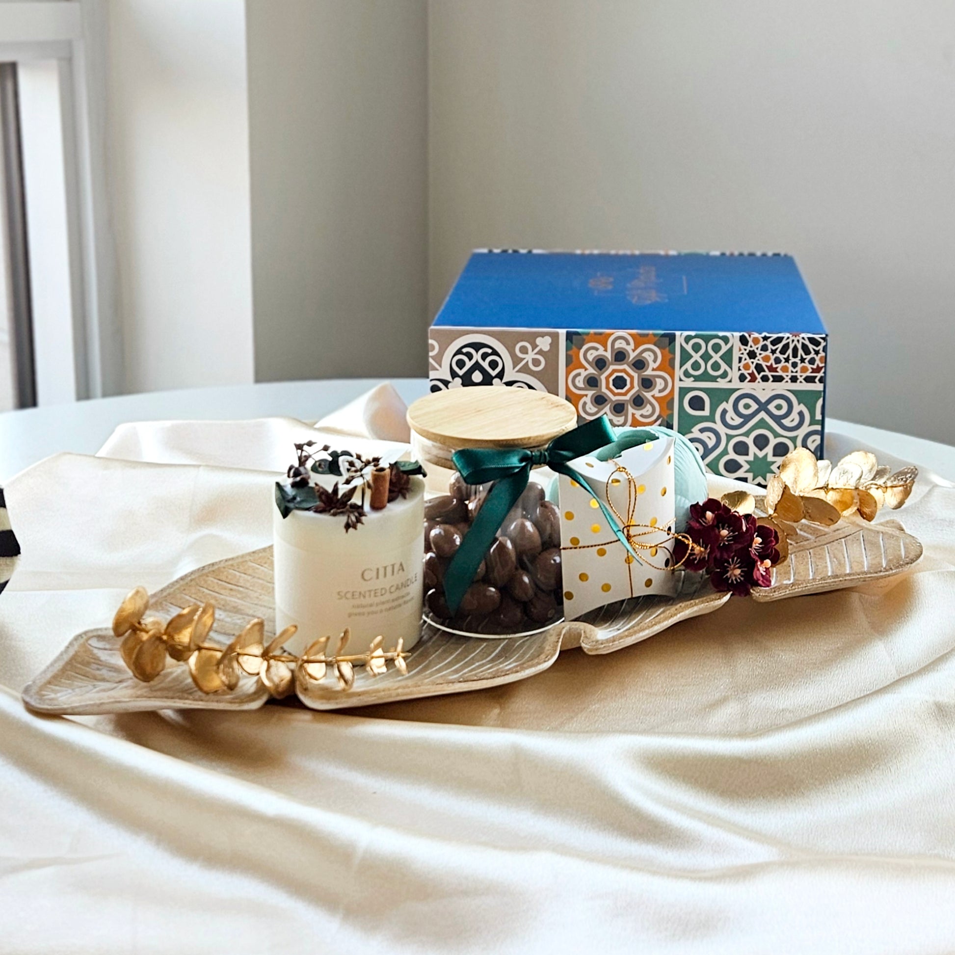 A gift set arrangement featuring a soy wax candle in a gold container, a rustic leaf tray, a blue and gold box with a decorative design, and a paper pocket with a gold polka dot pattern containing a pair of gold-coated chocolate almonds.