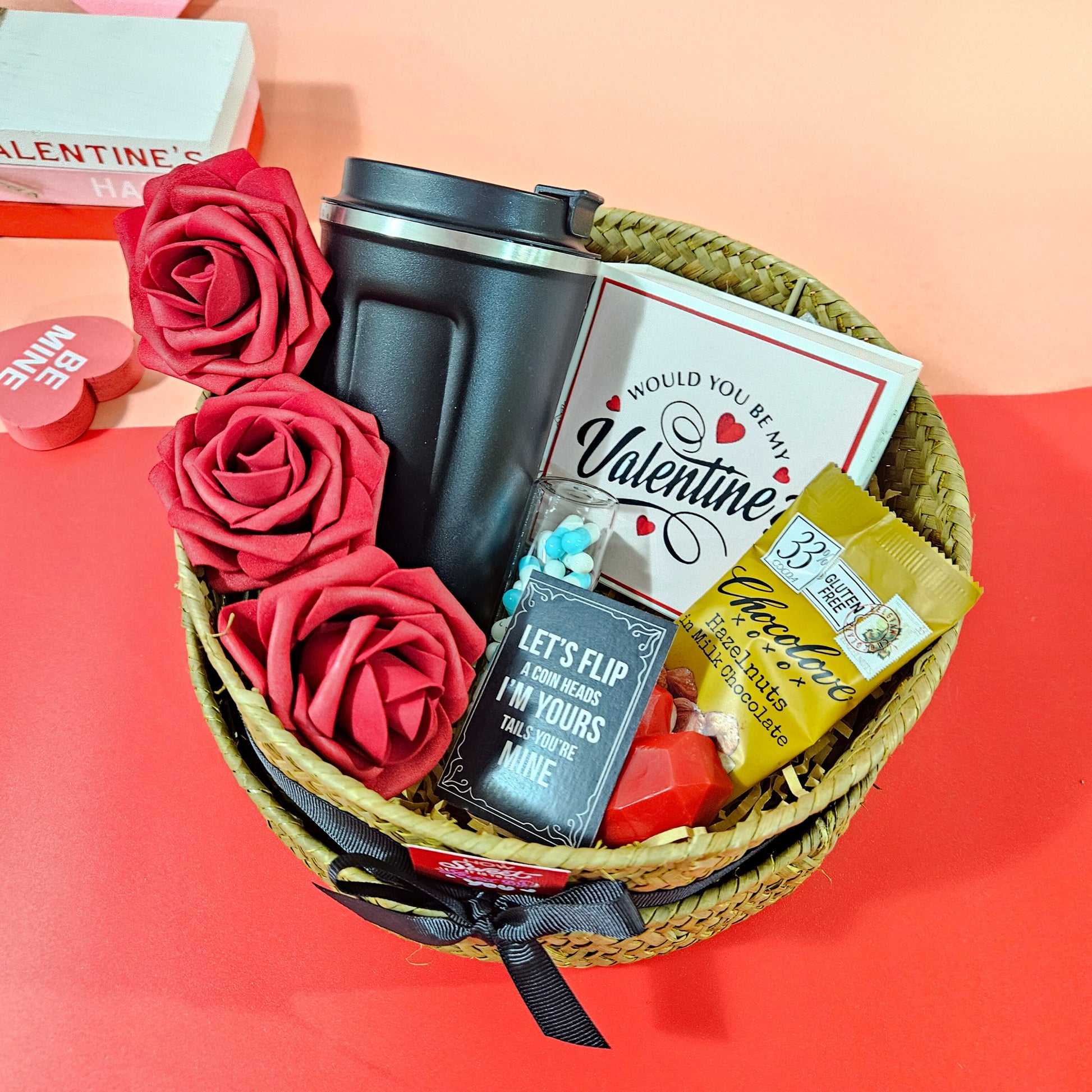 A Valentine's Day gift hamper basket containing a black airtight flask, red roses, a hazelnut chocolate bar, a heart-shaped scented candle, message capsules, and a soy wax car diffuser candle.