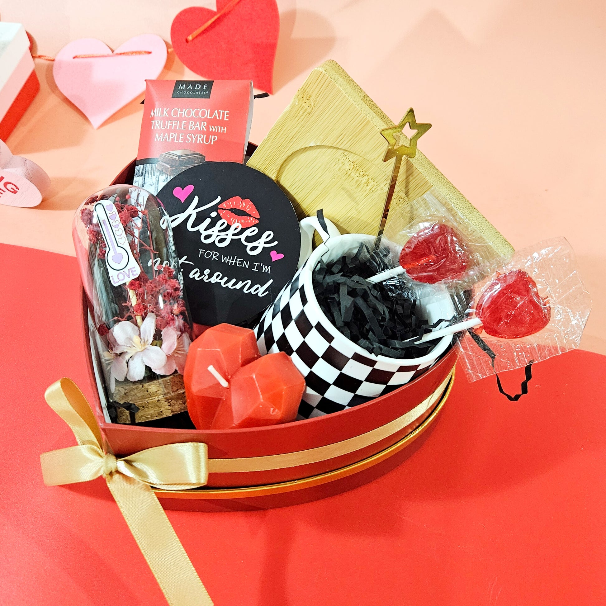 A Valentine's Day gift hamper containing a heart-shaped box, a checker coffee cup with wooden coasters, a heart-shaped candle, a miniature cloche with a personalized message, and a chocolate treat.