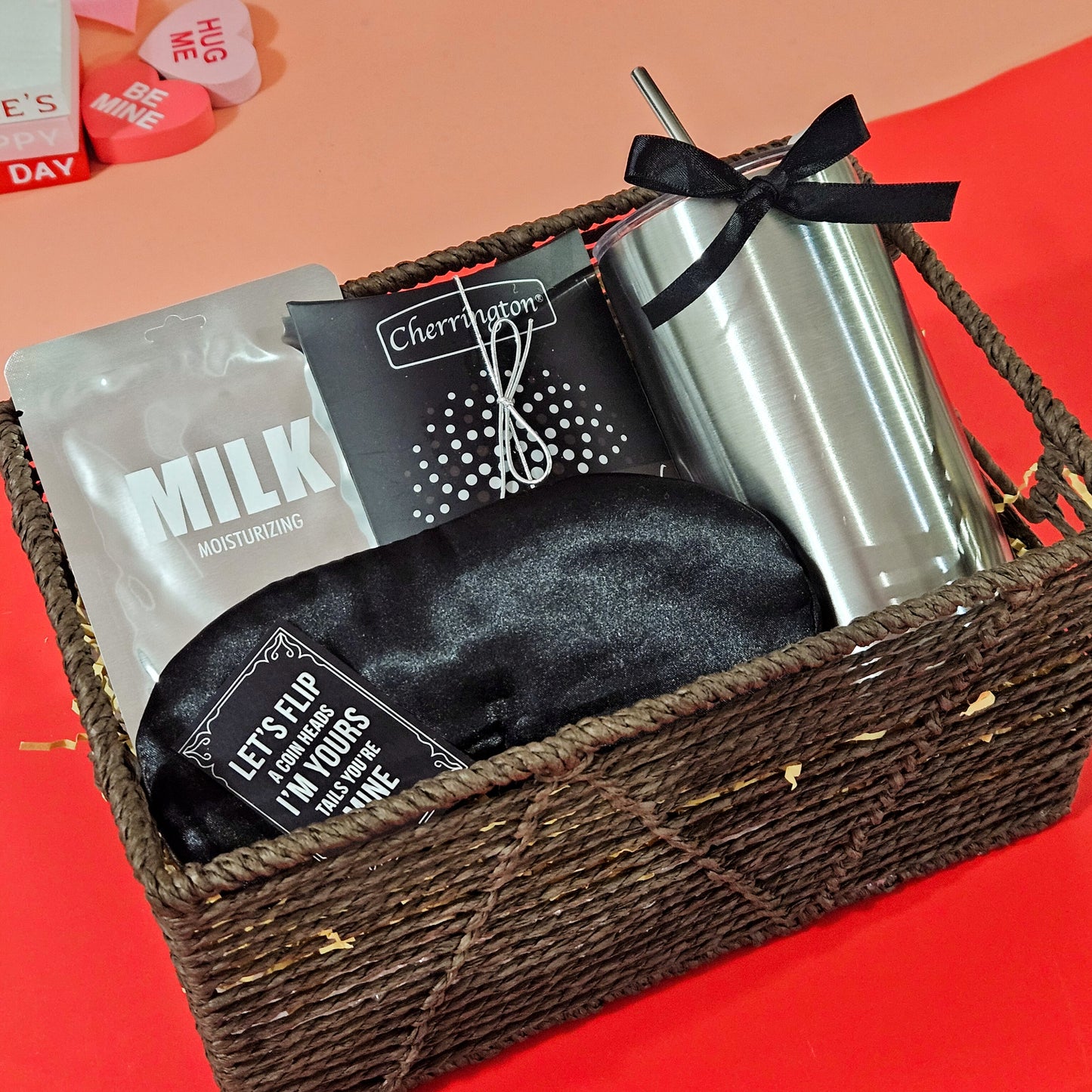 A gift basket containing a black satin eye mask, a 'Milk' face mask from LAPCOS, mint drops, and a stainless steel sipper, all arranged in a rustic brown thread basket.
