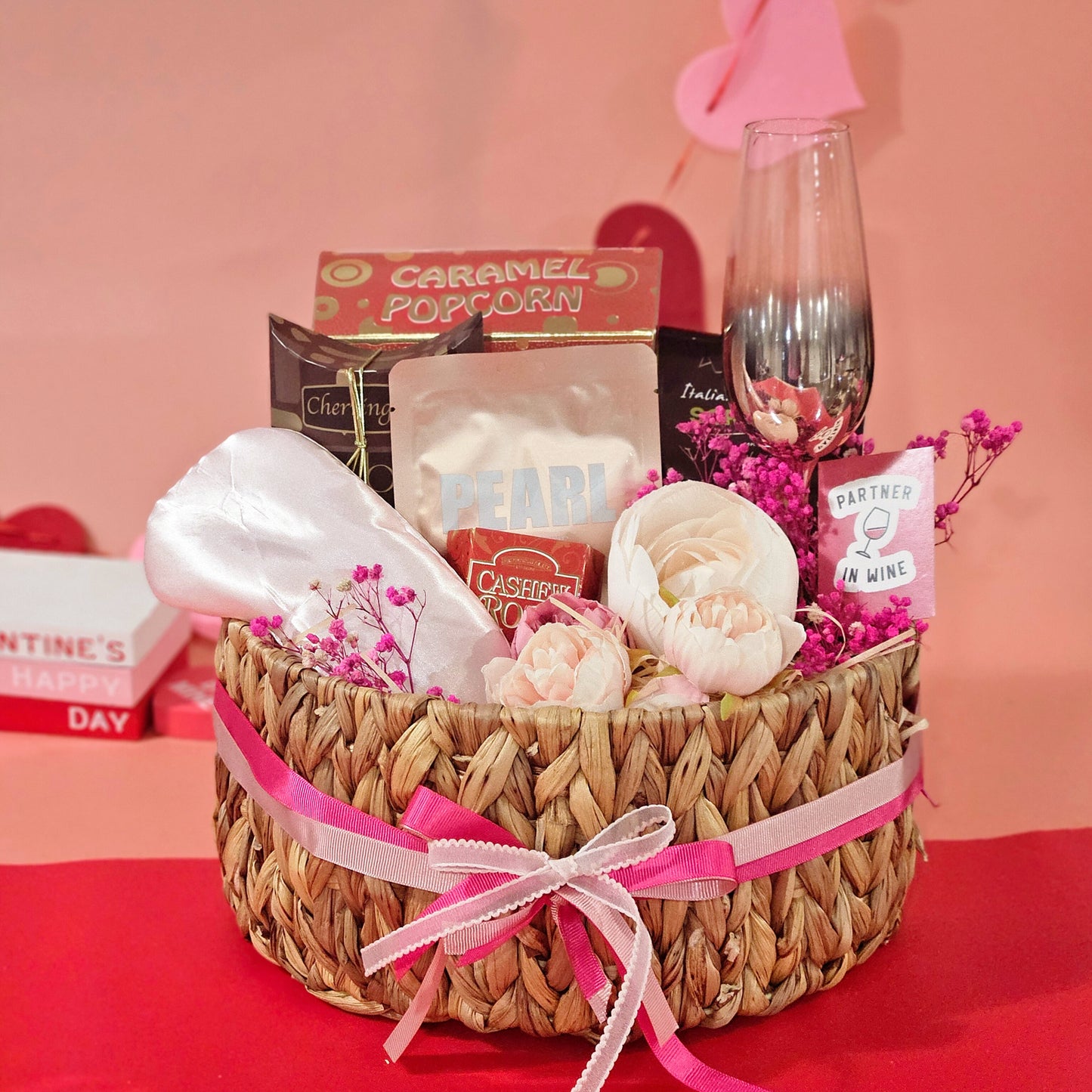 A wicker basket gift hamper arranged with a champagne glass, gourmet cracker, caramel popcorn, face mask, eye mask, chocolate drops, and almond cashew roca, all presented on a pink background.