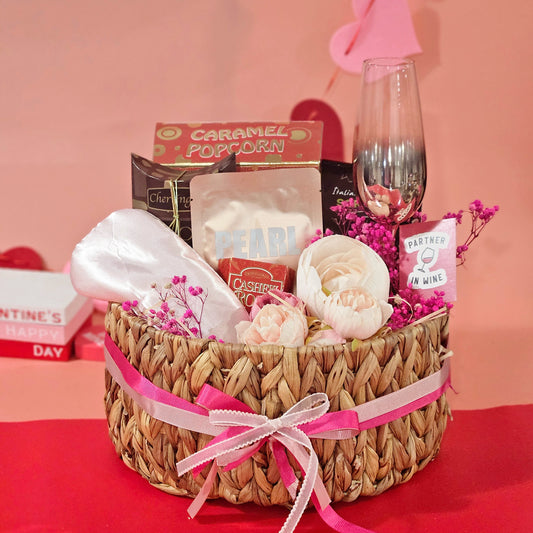 A wicker basket gift hamper arranged with a champagne glass, gourmet cracker, caramel popcorn, face mask, eye mask, chocolate drops, and almond cashew roca, all presented on a pink background.