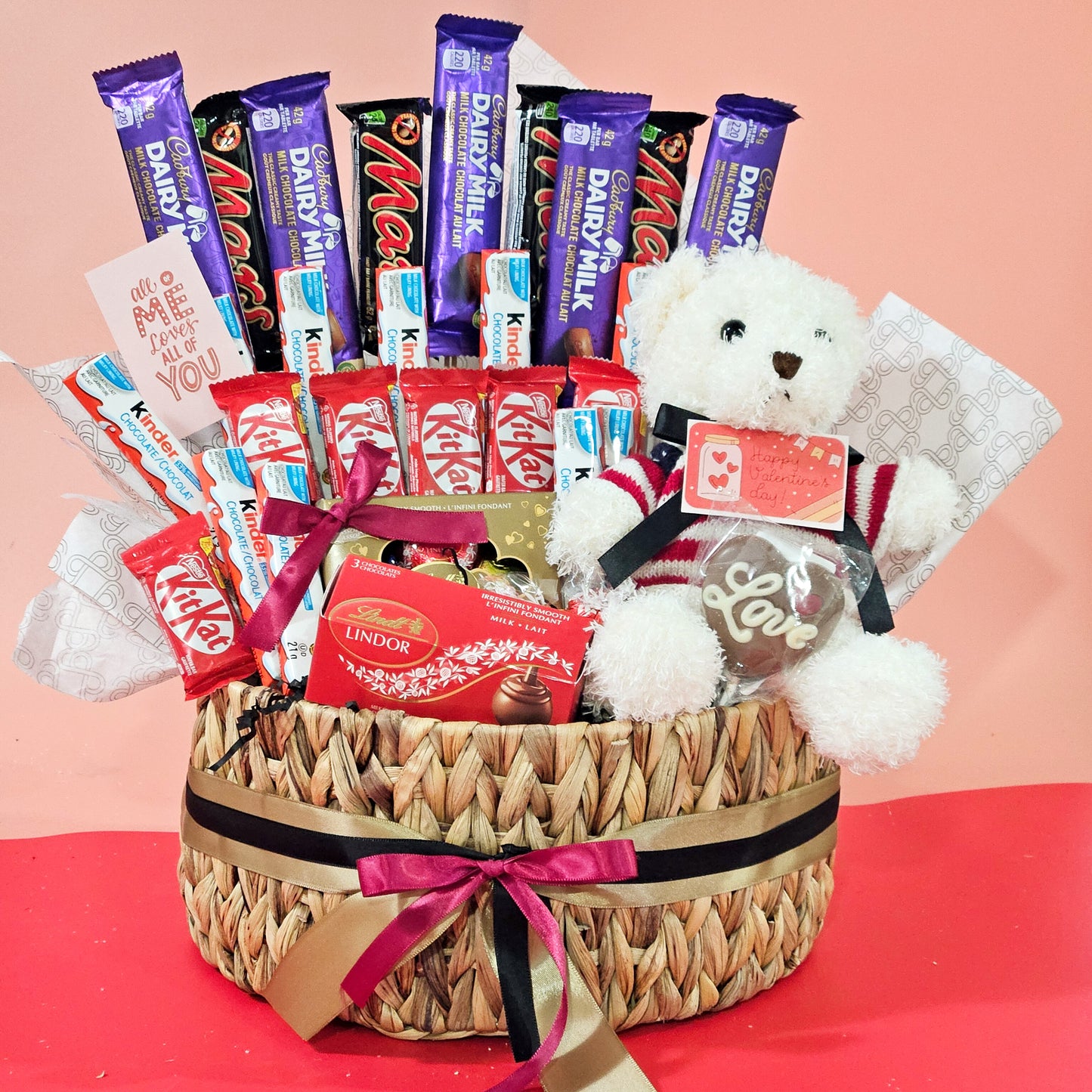 A wicker basket containing an assortment of chocolate bars and a white teddy bear, adorned with a red and white bow.