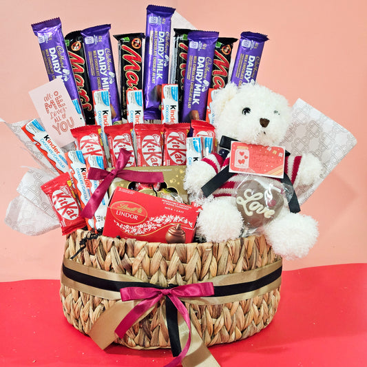A wicker basket containing an assortment of chocolate bars and a white teddy bear, adorned with a red and white bow.