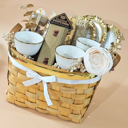 A wicker basket gift hamper arranged with white floral accents, including a set of white cups and saucers, gourmet popcorn, and other assorted gourmet treats.