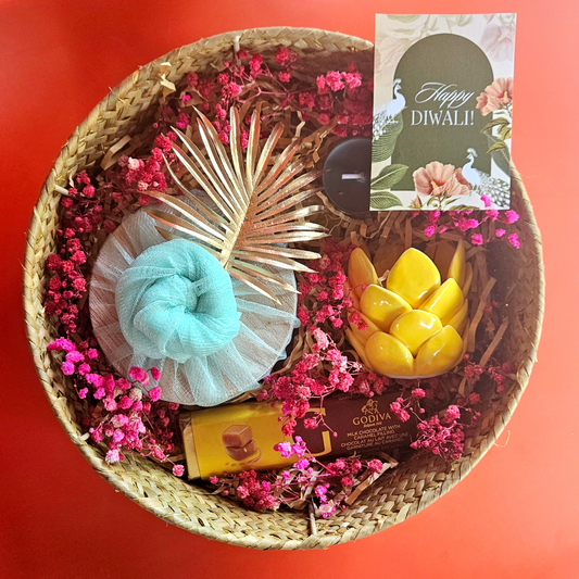 A wicker basket containing a green momo box, a yellow porcelain diya, a Godiva chocolate bar, and decorative flowers, with a 'Happy Diwali' greeting card.
