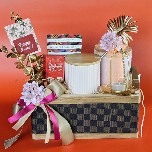 A brown checked basket with a gold-plated hanging lantern, a pastel coffee mug, a pack of almond-coated Roca, a mix of dry berries, a textured tin with Indian savory treats, and a red 'Happy Diwali' greeting card.