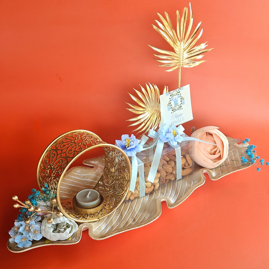 A decorative wooden banana leaf platter with various items including almonds, cashews, and a circular Indian diya, arranged on a red background.