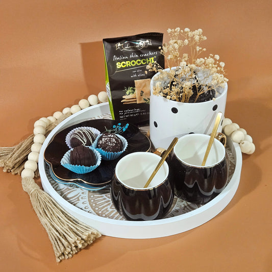 A gift hamper set with a Moroccan breakfast theme, including a round plate with wooden beads, scalloped cups and saucers, a planter, dates, and crackers presented on a tray.
