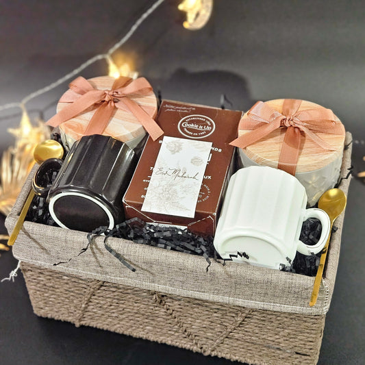 A gift basket containing a pair of white and black cups with saucers, gold stir spoons, a box of cookies, a tin of dates, and a tin of Turkish delights, all arranged in a woven basket with decorative lights in the background.
