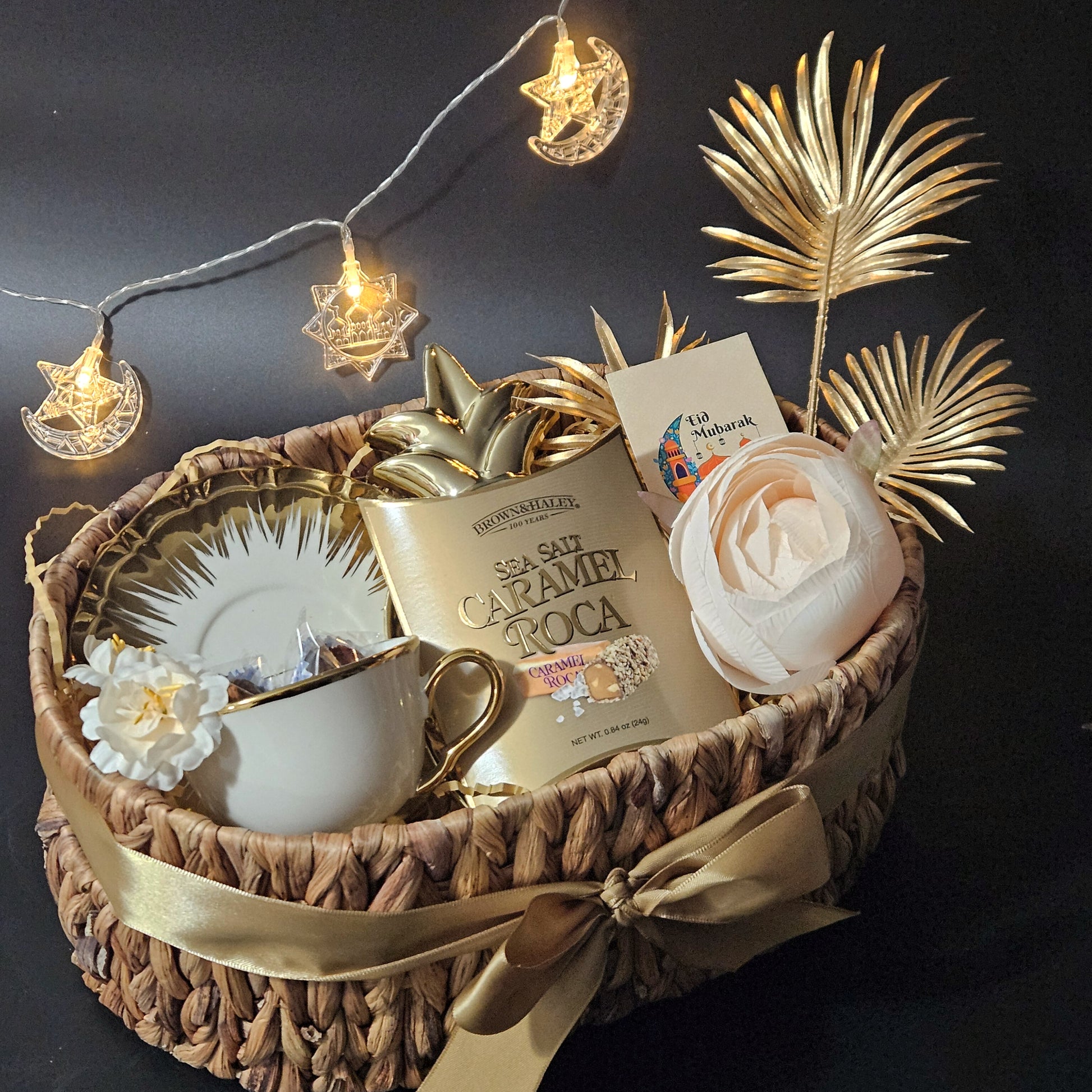 A festive hamper gift basket with an oval-shaped cane basket, containing a white gold-plated queen cup and saucer, a pineapple-shaped trinket, a pair of stuffed dates, and caramel Roca.