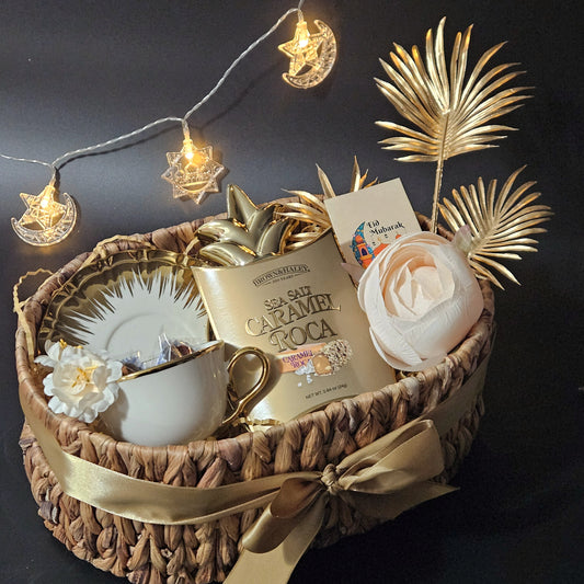 A festive hamper gift basket with an oval-shaped cane basket, containing a white gold-plated queen cup and saucer, a pineapple-shaped trinket, a pair of stuffed dates, and caramel Roca.
