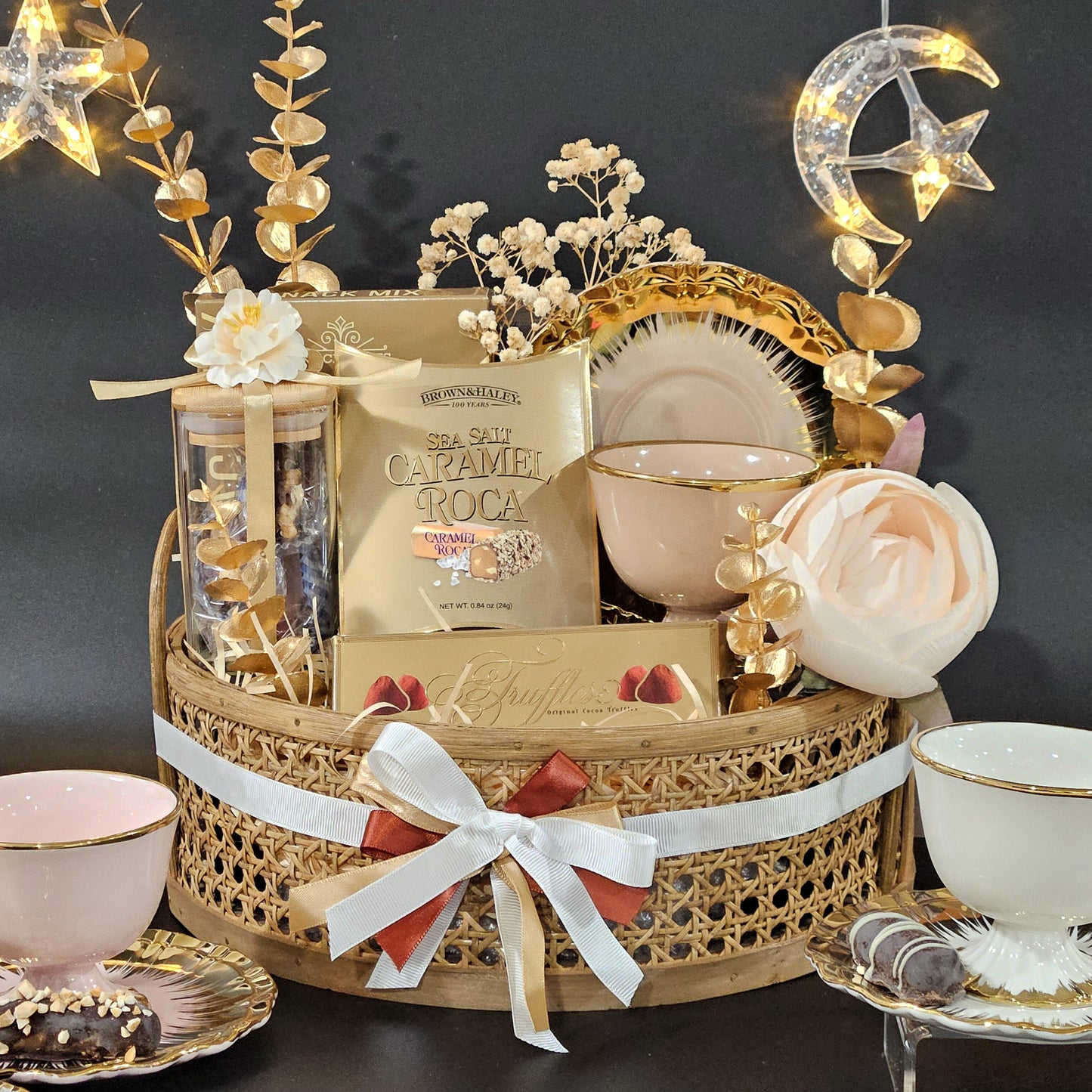 A festive gift basket with a golden rattan base, featuring a Queen's Cup & Saucer with gold embellishments, a jar of nuts, and gourmet cookies, all tied together with a white ribbon.
