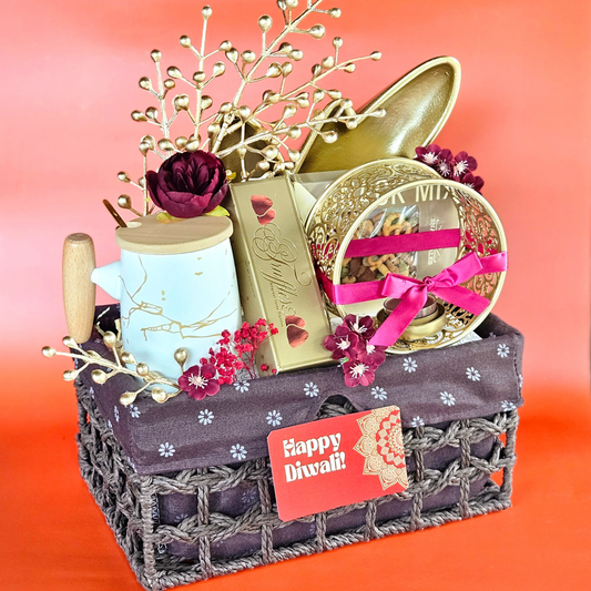 A weaved metal basket with a red and gold color scheme, containing a white marble coffee mug, gourmet truffles, a pack of snack mix, a gold leaf platter, and a circular filigree work diya, with decorative flowers and a 'Happy Diwali' sign.