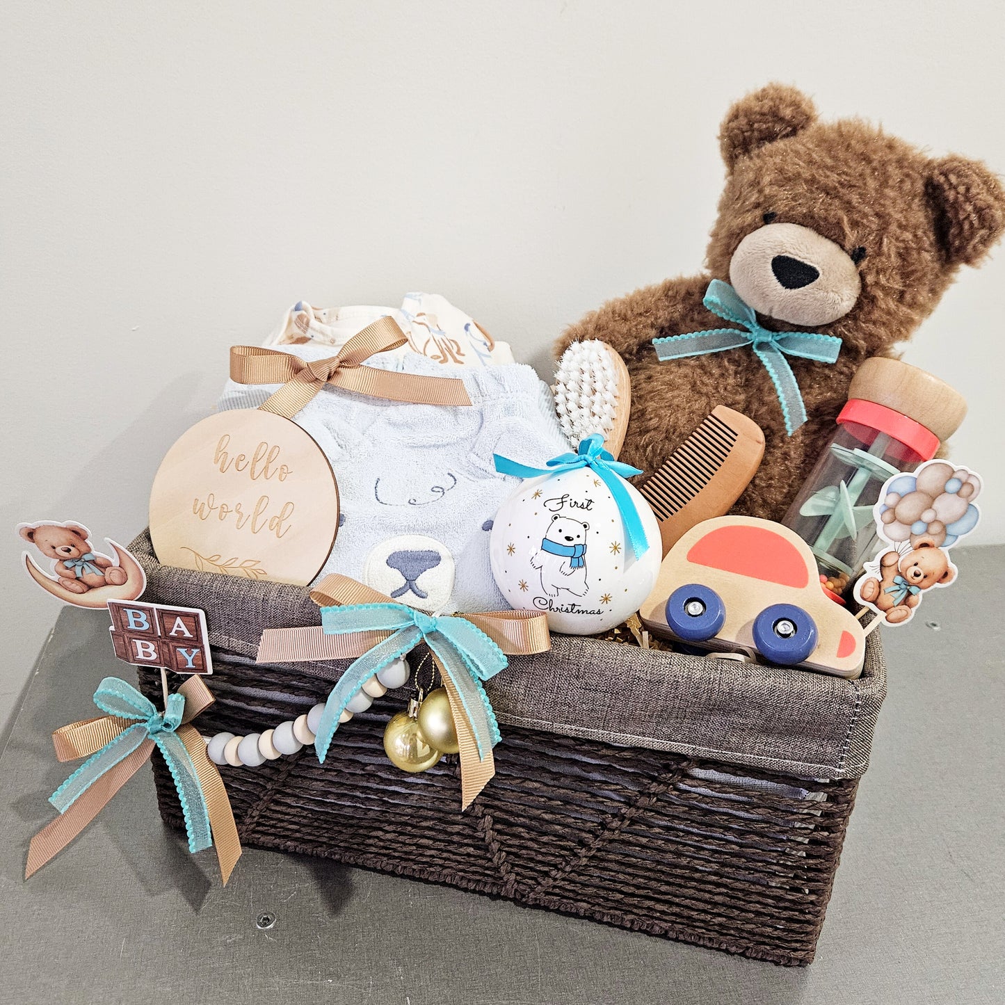 A wicker basket containing a teddy bear, a baby rattle, a pair of baby shoes, a wooden toy, a comb and brush set, and a few decorative items.