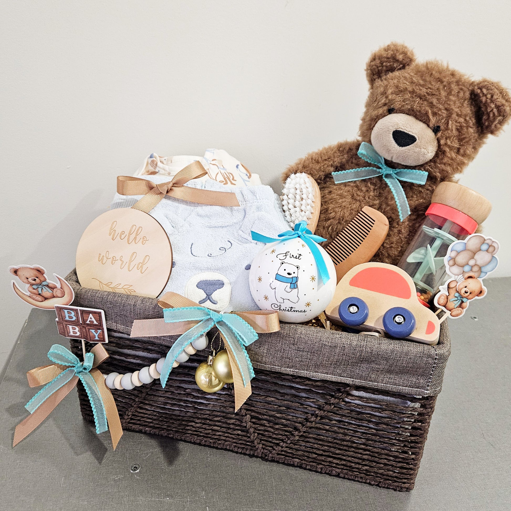 A wicker basket containing a teddy bear, a baby rattle, a pair of baby shoes, a wooden toy, a comb and brush set, and a few decorative items.