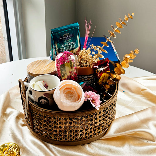 A rattan basket gift hamper containing a variety of items including a printed vase, a black tea cup, a tin with cookies, and a glass cloche with 'Eid Mubarak' plating, all beautifully arranged on a table with a golden yellow tablecloth.