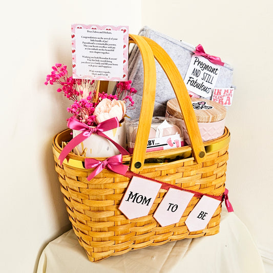 A gold rim picnic basket filled with various items for expectant mothers, including a bouquet of artificial flowers, a tin container with cookies, and a textured card with 'Mom to be' inscription.