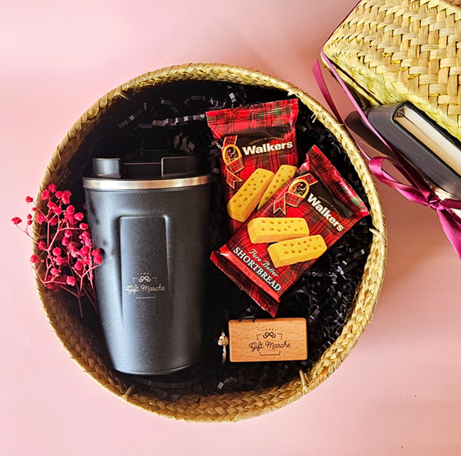 A gift set containing a black coffee flask, a pack of shortbread cookies, a wooden keychain, and a notebook, all presented in a woven round box.