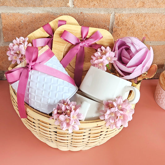 A wicker basket gift hamper with pink accents, including Turkish delight, a white cup, wooden coaster, and calla lily flowers.