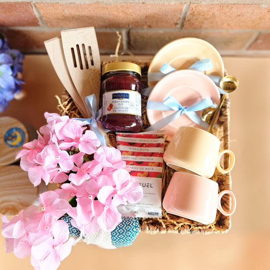 A square cane basket gift hamper filled with kitchen items including a pair of pink cups and saucers, wooden utensils, a pack of nuts, strawberry jam, and decorated with hydrangea flowers.