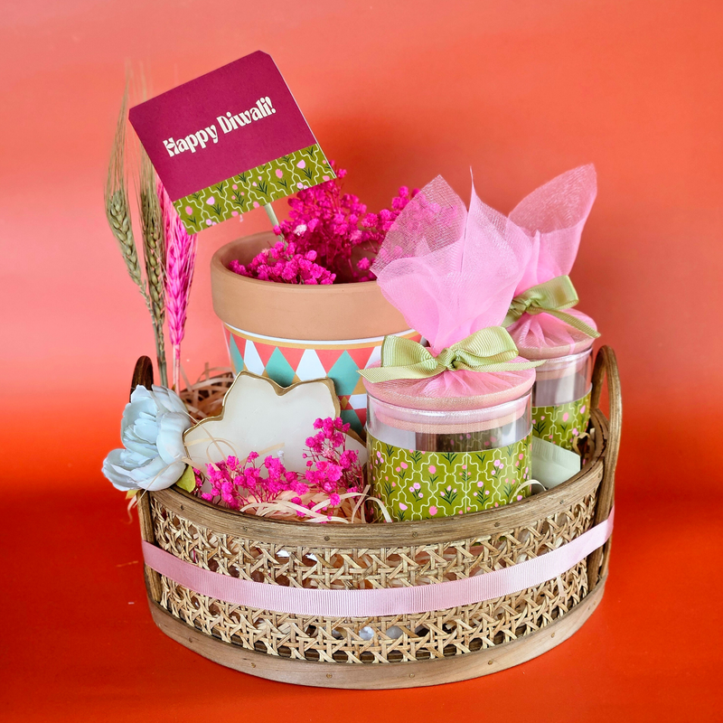 A Diwali hamper basket containing various items including two airtight containers, a printed planter, a gold plated leaf shape diya, and decorative flowers, with a 'Happy Diwali' card and pink tissue paper.