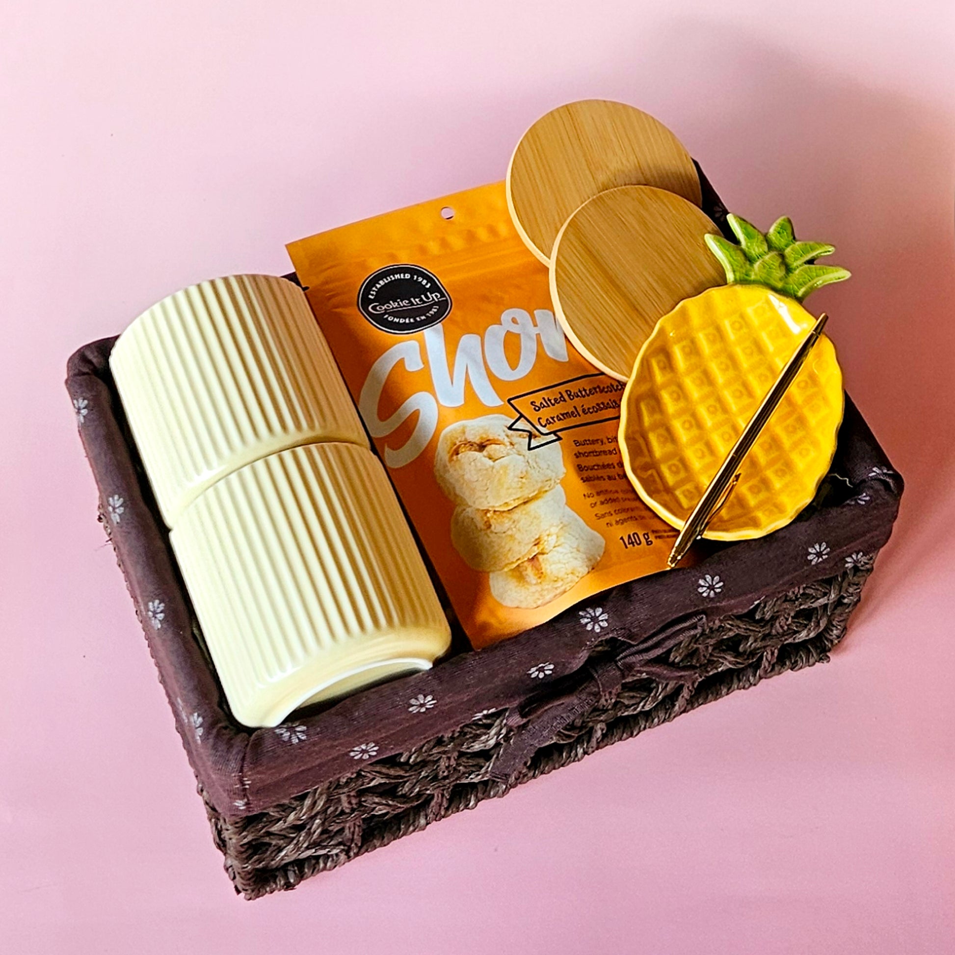 A corporate hamper gift basket containing a pair of white coffee mugs with wooden lids, a gold pen, and a pack of salted caramel cookies. The basket is wicker-like and presented with a pineapple trinket and a 'Quirky Hamper' branding card visible.