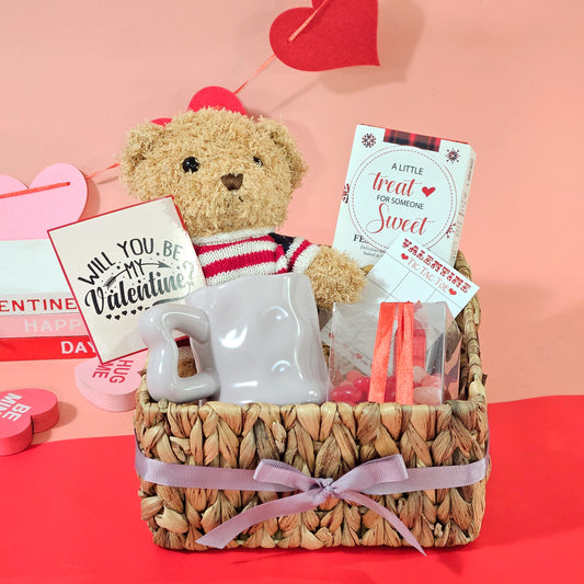 A Valentine's Day themed gift basket containing a teddy bear, a white coffee mug with hearts, gourmet cookies, and heart-shaped cinnamon gummies, all arranged in a square wicker basket with a pink ribbon.