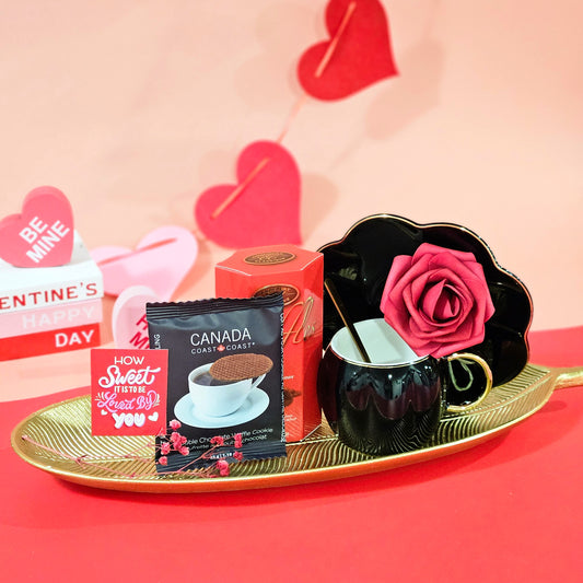 A luxurious gift hamper set on a red surface, featuring a gold leaf tray, black Scallop cup and saucer, a bar of truffle chocolate, and a premium waffle pack. Decorative hearts and a rose add to the elegant presentation.