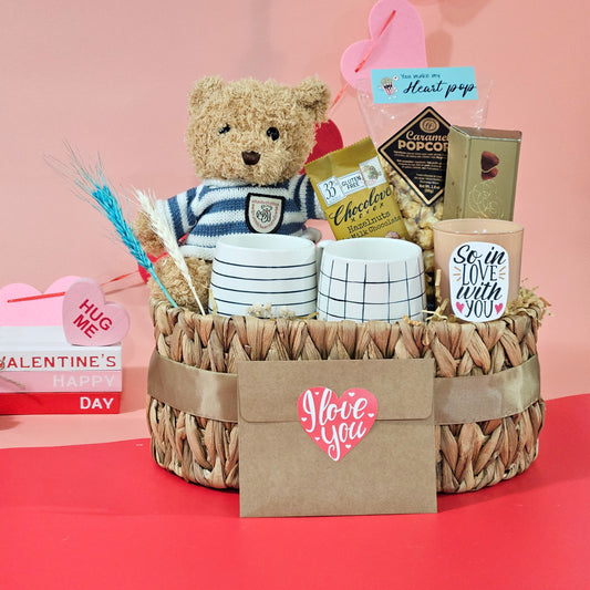 A Valentine's Day hamper basket containing a pair of navy blue coffee mugs with saucers, a brown mini teddy bear, caramel popcorn, truffle and hazelnut chocolates, and a scented candle, all arranged in an oval-shaped wicker basket with a 'I love you' card in front.
